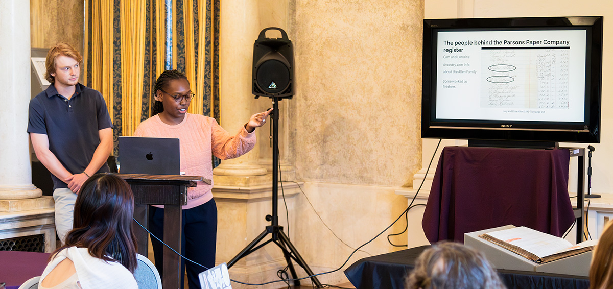 Two students at a podium pointing at a screen with text and data on it