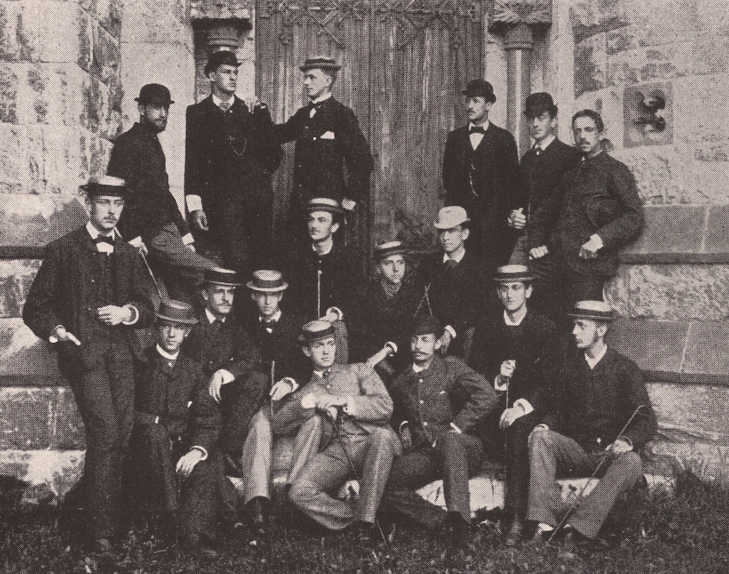 A black and white photo of young men in suits against a brick wall