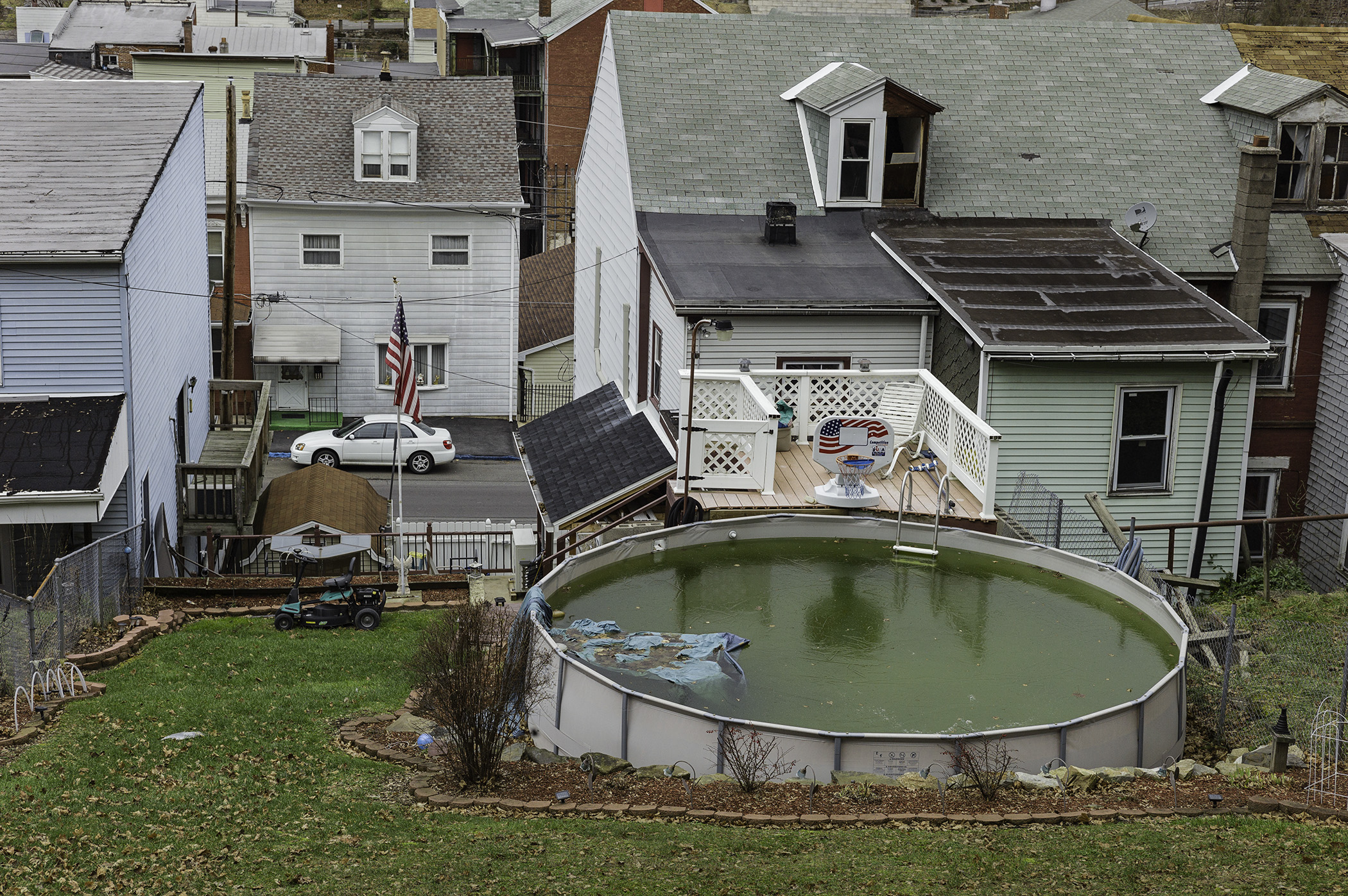 Photograph taken from the top of a hill in a suburban neighborhood, an above-ground backyard pool with green water and a blue fl