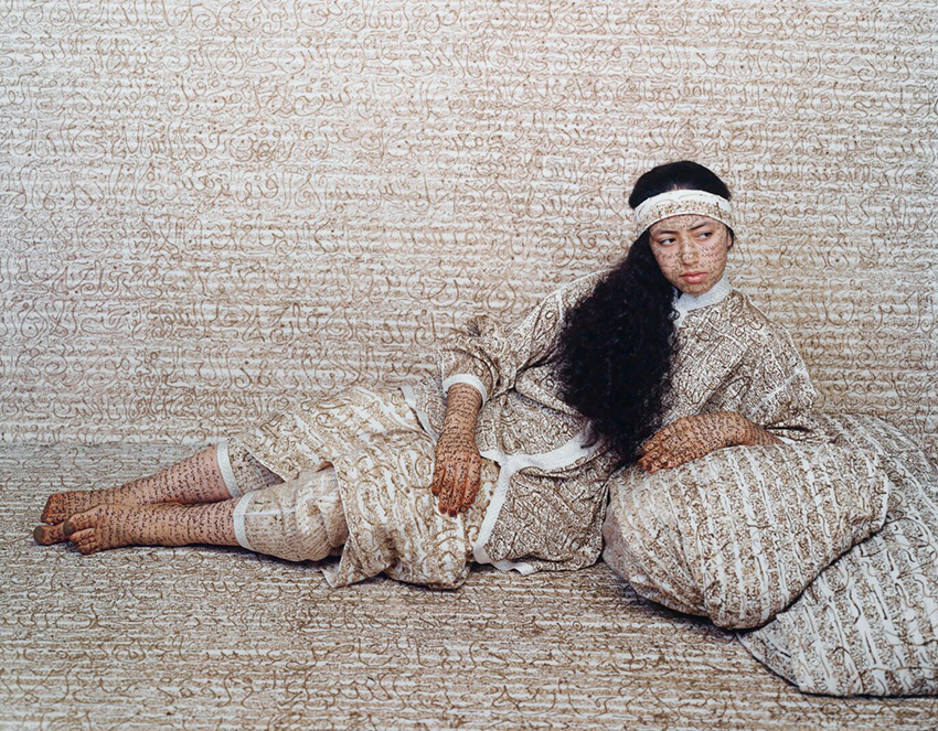 Photograph by Lalla Essaydi of a woman lying with elbow propped on pillow, surrounded by calligraphy text