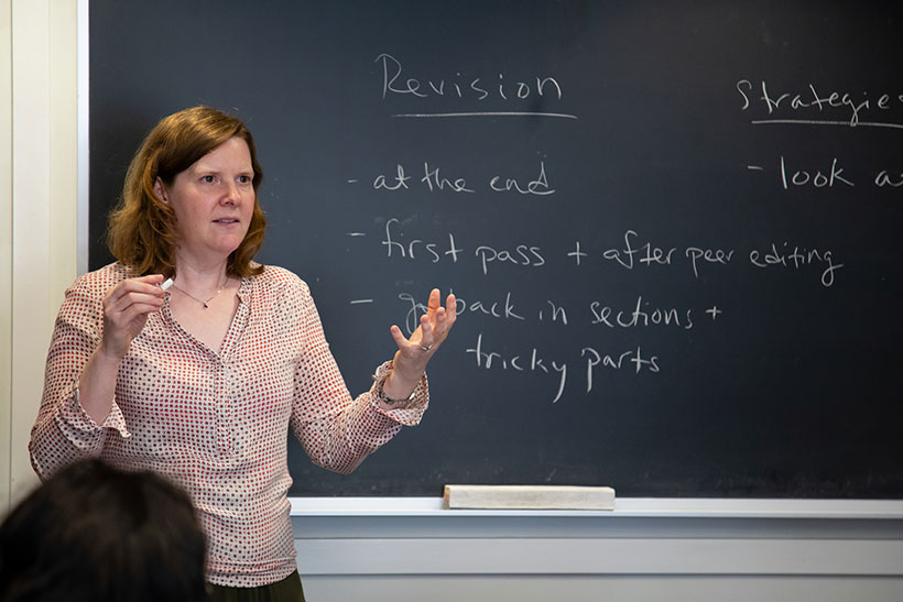 Professor Sara Brenneis stands before a blackboard covered with writing, speaking to her students