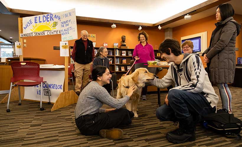 Two students sit on the floor petting a golden retriever. In the background is the Wise Elders Advice booth is visible