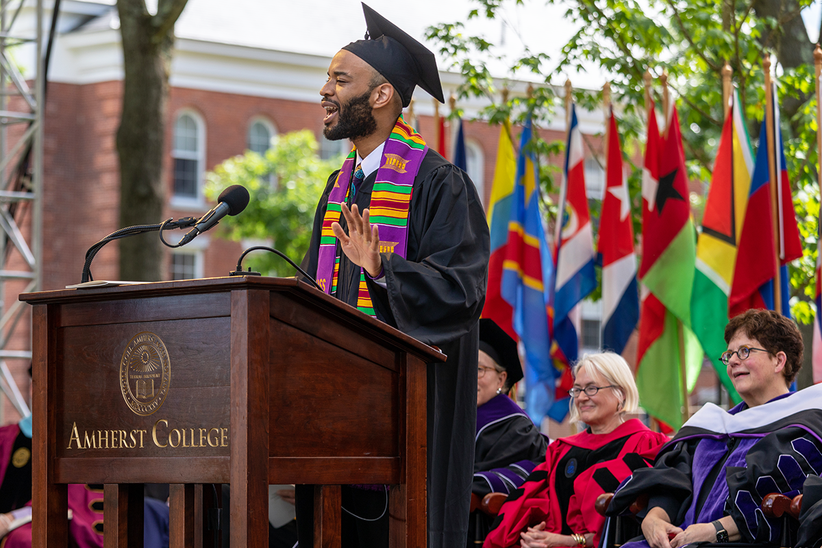 A young man in commencement regalia giving a speech at a podium that says Amherst College