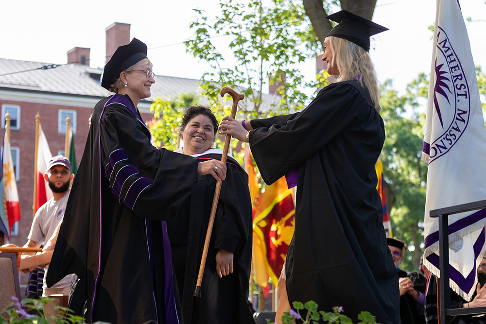 A woman handing another woman a cane, both wearing commencement regalia