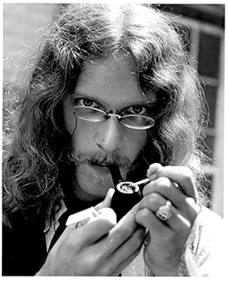 A bearded young man with long hair smoking a pipe 