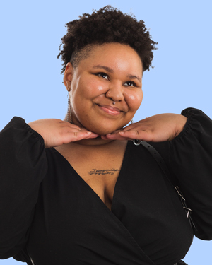 Ash Smith, a fat, black transmasculine person with facial piercings smiles while supporting his chin with his hands