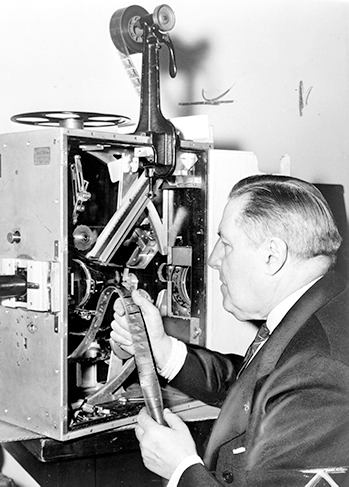 A black and white photo of a man inspecting film coming out of a projector