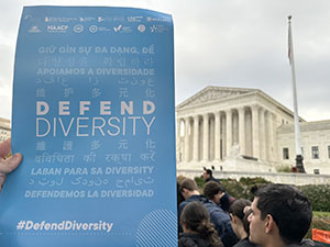 A hand holds up a Defend Diversity site in front of the US Supreme Court building