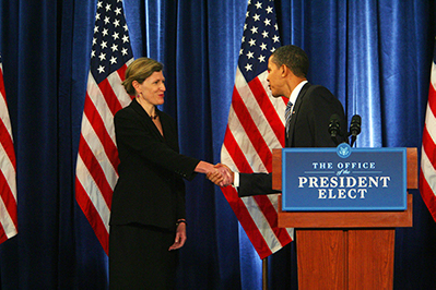 A photo of Jeanne Lambrew shaking hands with President Obama in front of a series of American flags