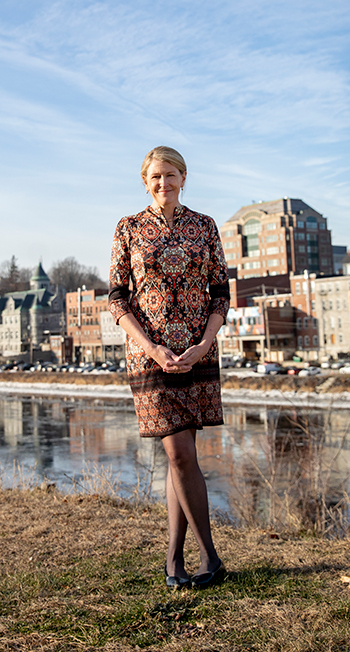 A woman in a colorful dress standing in front of a river with buildings and houses in the distance.