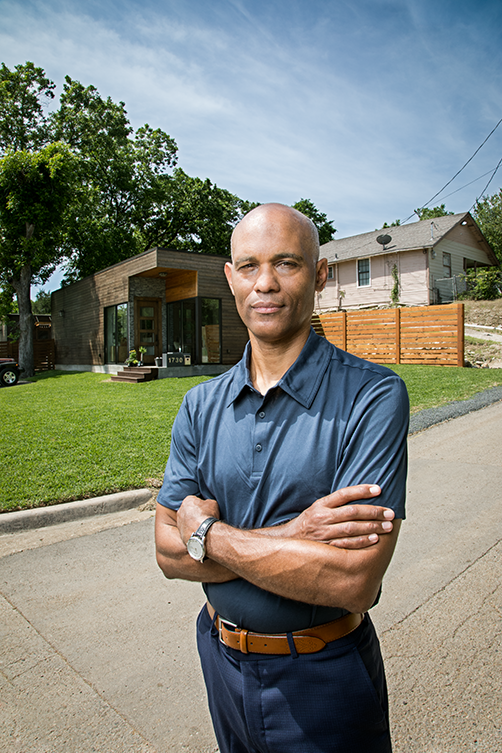 A photo of a Black man standing in front of a modern looking house with his arms crossed