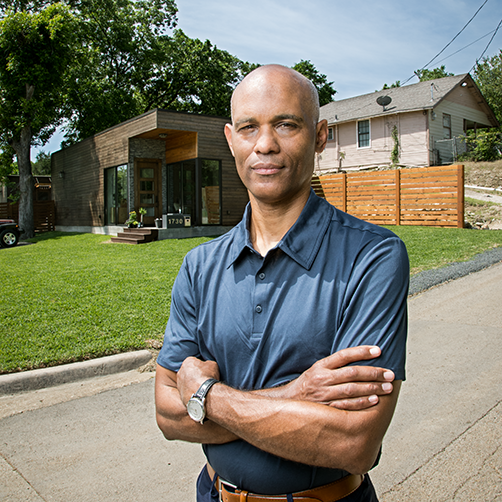 A Black man with his arms folded standing in front of a modern house