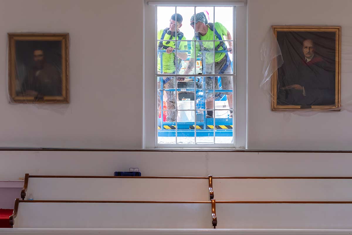 Two workers applying protective paint to the outside of a window in Johnson Chapel.