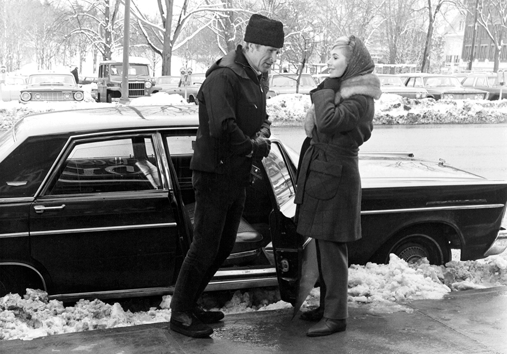 A black and white movie still of a man and woman standing next to a car with the door open