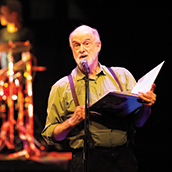 An older man on stage reading from a large book