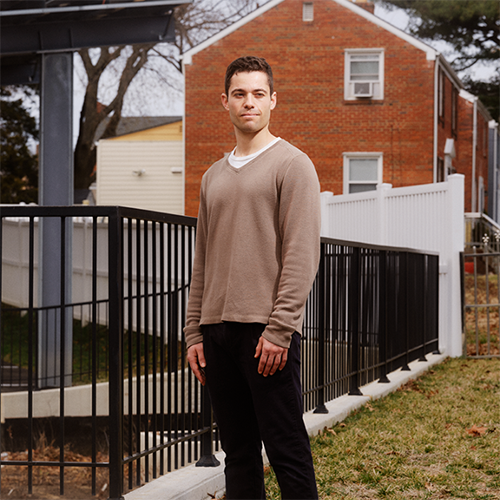 A man in a brown sweater standing in front of a fence