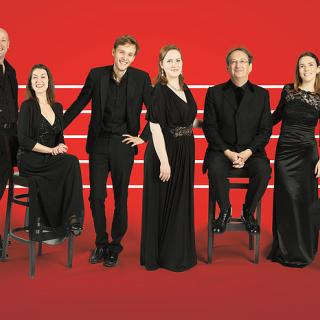 11 Tallis Scholars dressed in black, lined up in front of a musical staff in front of a red background 