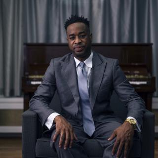 Damien Sneed seated in an armchair in front of a piano, wearing a suit and tie