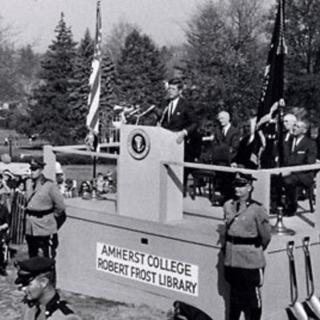 Black-and-white image of Kennedy speaking at a raised podium on the Amherst College campus, surrounded by guards and spectators