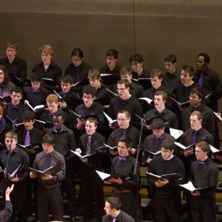 Concert Choir standing on stage, dressed in black, with Mallorie Chernin conducting