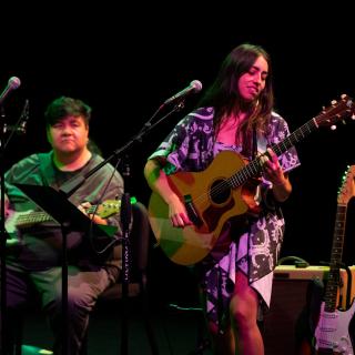 Nokosee Fields playing guitar onstage with Mali Obamsawin, who is playing guitar and singing