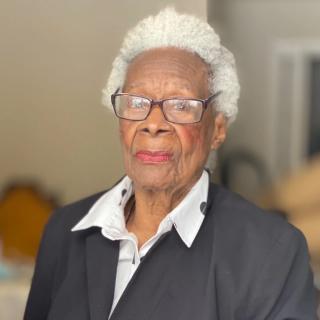 Annye C. Anderson with short white hair, glasses, a white collar and black jacket, looks unsmiling into the camera