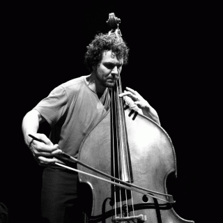 Black and white photo of the top half of a middle-aged white man in a T-shirt with tousled hair, pulling a bow across the strings of a double bass, as he holds down one finger on the fret-board