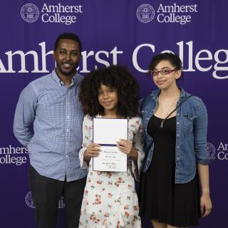 An award recipient and two others stand in front of an Amherst media backdrop