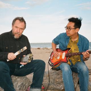 The two members of 75 Dollar Bill sitting on rocks on a beach, holding musical instruments