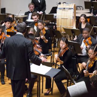Violinists, drummers and other musicians from the Amherst Symphony Orchestra playing onstage in Buckley Recital Hall, being conducted by Mark Lane Swanson