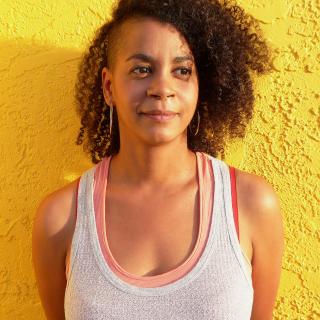 Aisha Sabatini Sloan wearing layered tank tops and hoop earrings, standing in front of a yellow wall
