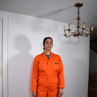 Image of a barefoot woman in an orange jumpsuit. A brown chandelier hangs from the ceiling and casts a shadow on a blank white wall. 
