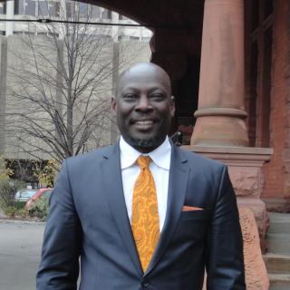 Closeup of Ato Quayson standing outdoors, wearing a suit and tie and smiling