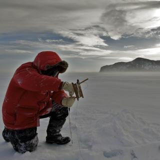 Davila research image showing a researcher wearing a read parka and kneeling down amid a snowy landscape, under a cloudy sky