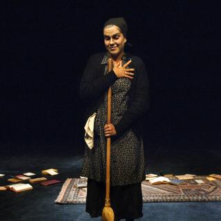 A woman standing on a dark stage holding a broom, with a rug and books scattered behind her