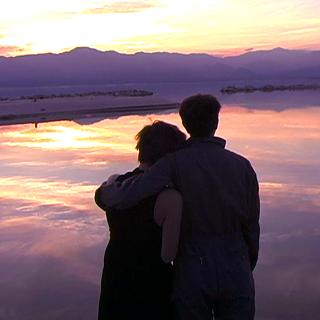 Image of one person with their arm around another as they look out over a body of water toward mountains at either sunrise or sunset