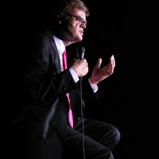 Color image of Garrison Keillor sitting on a stool and holding a microphone