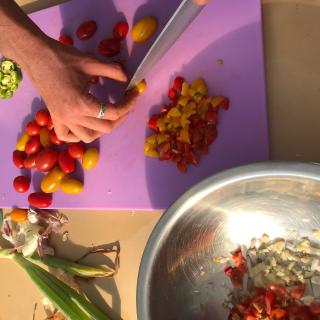 Hand chopping cherry tomatoes on a cutting board with a bowl of garlic nearby.