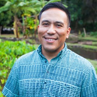 Photograph of Ty P. Kāwika Tengan. He is wearing a blue shirt with a turquoise pattern. He is smiling and standing in a patch of bright green leaves.  He has short dark hair.