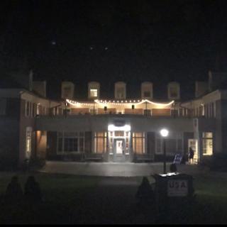 Image shows the Valentine Balcony lit up by string lights at night, some students sitting in the grass on the quad looking at the balcony where music is being played.