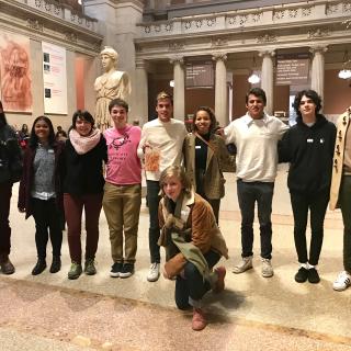 Nine students standing in a row inside a New York City art museum, with a 10th student kneeling in front of them