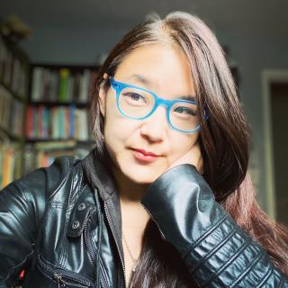 Janice Lee wearing a black leather jacket and blue-framed eyeglasses. Bookcases and part of a doorframe are visible in the room behind her.