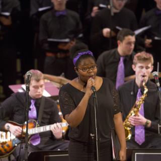 A young woman singing into a microphone, with several jazz musicians playing behind her