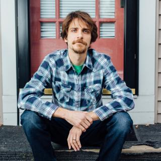 Joseph Scapellato seated on the front steps of a house, wearing a plaid shirt and blue jeans