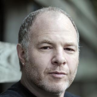 this is a photo of Jackson Katz.  He is a white man with a shirt few day growth beard, with brown hair with grey.  he is staring at the camera, wearing a serious expression. he is wearing a black shirt and the background is blurry.