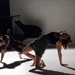 Two dancers contorting on a studio floor, in front of a grand piano and other musical instruments