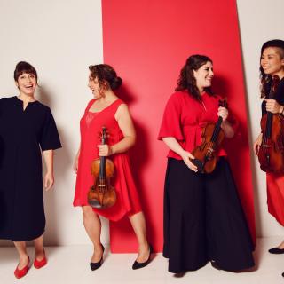 The four members of the Aizuri Quartet dressed in red and black, standing in front of a red and white wall, holding their respective instruments and smiling at one another