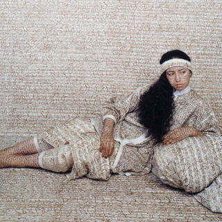 A young woman with long dark hair, lounging on pillows; her clothing and skin, as well as the pillows and background, are covered in swirling marks that may be calligraphy