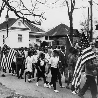 Black-and-white photo of a crowd marching with U.S. flags past houses and trees