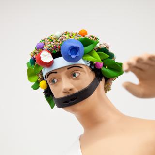 Image of the head, neck, shoulders and raised left hand of a human figure with a colorful floral headdress and a black strap across his mouth